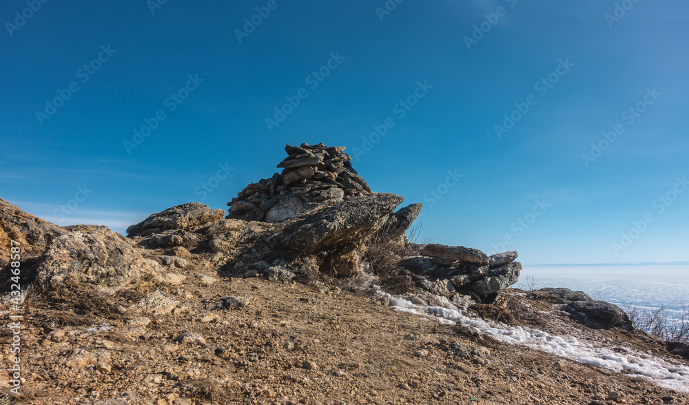 Picturesque cliffs, devoid of vegetation, against the backdrop of a clear blue sky. Cracks on the stones. There is some snow on the ground. The surface of the frozen lake is visible. Baikal
