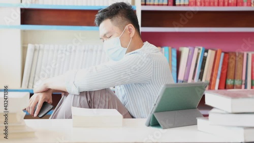 A young Asian man wearing a mask in his office is sitting in exhaustion and holding his head in exhaustion, the concept of spacing and detention and working at home during the coronavirus outbreak.