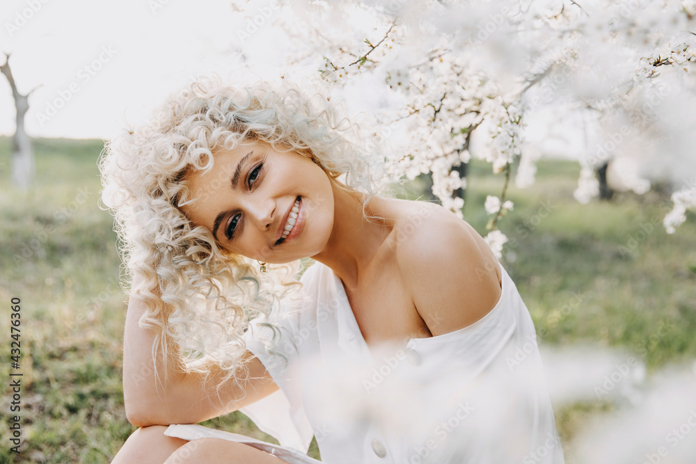 Young blonde woman with curly hair, wearing a white dress, sitting under a cherry tree in bloom, smiling. Young blonde woman with curly hair, wearing a white dress, sitting under a cherry tree in bloom, smiling.
