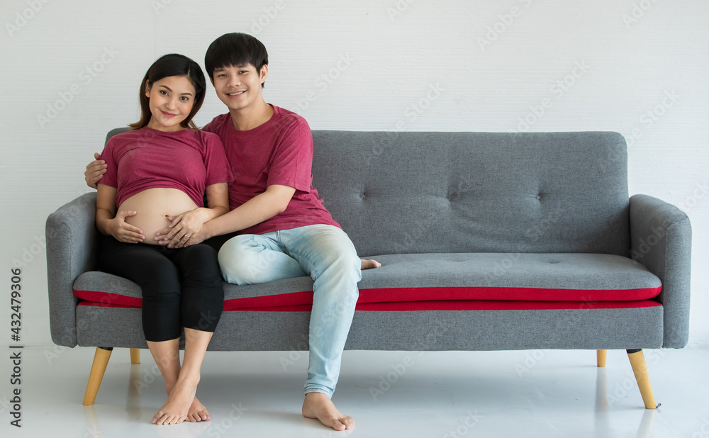 young family husband and wife sitting on a couch. They are smiling and looking at a camera. A man and pregnant woman touching on a belly. Expecting a healthy baby young family husband and wife sitting on a couch. They are smiling and looking at a camera. A man and pregnant woman touching on a belly. Expecting a healthy baby