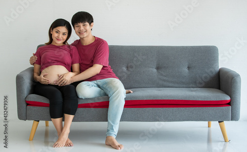 young family husband and wife sitting on a couch. They are smiling and looking at a camera. A man and pregnant woman touching on a belly. Expecting a healthy baby