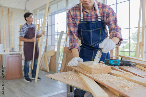 Young asian male worker wearing plaid shirt and blue jean apron with white gloves holding saw cutting wood stick on the table full of building tools and equipments while his colleague stand holding 