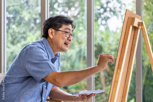 Senior smiling Asian man using brush and water color to paint on canvas. Happy retirement and activity concept.