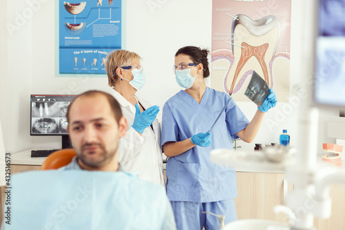 Stomatologist doctor and orthodontist assistant examining teeth radiography checking somatology problem while sick patient waiting on dental chair. Man preparing for surgery appointment