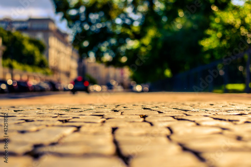 Summer in the city, the sunlit empty old street paved with stone next to the road on which cars are traveling. Close up view from the level of paving stones