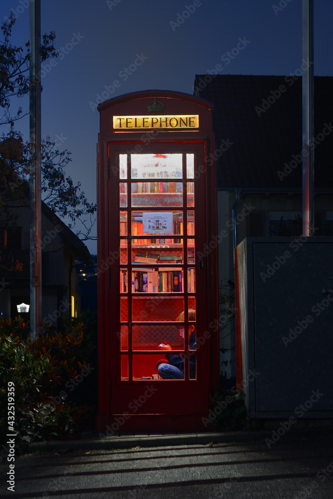 Fototapeta premium Boy reading at night in a red telephone box filled with books