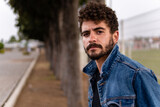 Handsome young man with curly hair and beard wearing denim posing in a public park. Close up portrait with confident expression and copy space