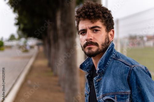 Fotografia, Obraz Handsome young man with curly hair and beard wearing denim posing in a public park