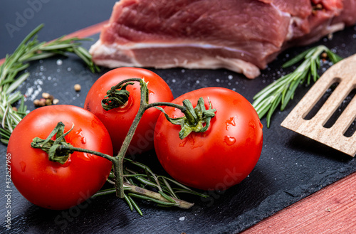 Fresh pork meat with ingredients for cooking on dark background. Tomato and rosemary