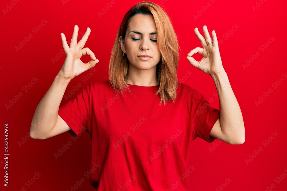 Hispanic young woman wearing casual red t shirt relax and smiling with eyes closed doing meditation gesture with fingers. yoga concept. Hispanic young woman wearing casual red t shirt relax and smiling with eyes closed doing meditation gesture with fingers. yoga concept.