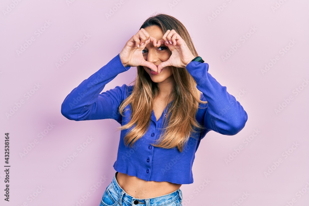 Beautiful hispanic woman wearing casual blue shirt doing heart shape with hand and fingers smiling looking through sign