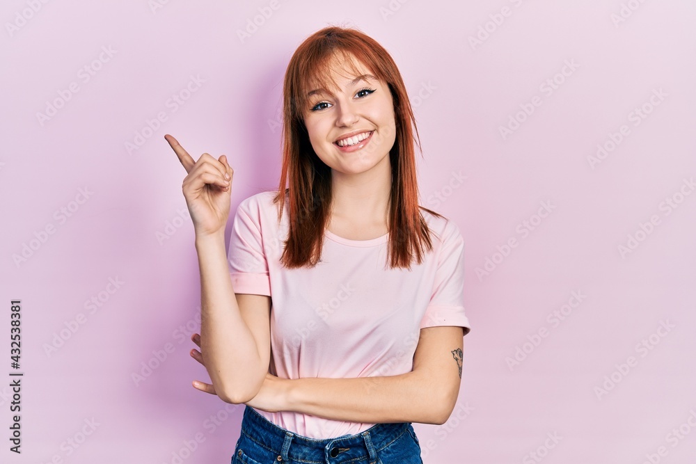 Redhead young woman wearing casual pink t shirt with a big smile on face, pointing with hand and finger to the side looking at the camera. Redhead young woman wearing casual pink t shirt with a big smile on face, pointing with hand and finger to the side looking at the camera.