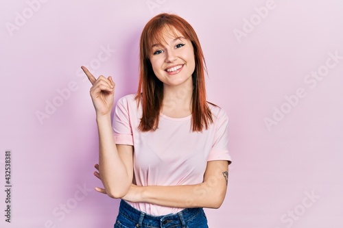 Redhead young woman wearing casual pink t shirt with a big smile on face, pointing with hand and finger to the side looking at the camera. © Krakenimages.com