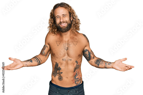 Handsome man with beard and long hair standing shirtless showing tattoos smiling showing both hands open palms, presenting and advertising comparison and balance