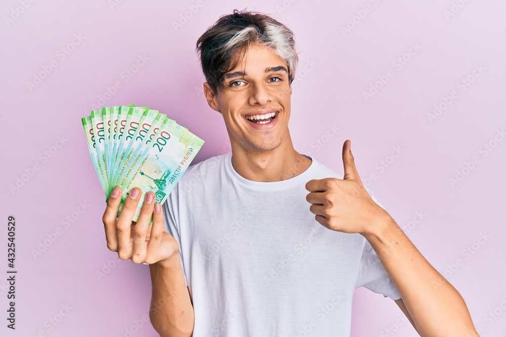 Young hispanic man holding russian 200 ruble banknotes smiling happy and positive, thumb up doing excellent and approval sign Young hispanic man holding russian 200 ruble banknotes smiling happy and positive, thumb up doing excellent and approval sign