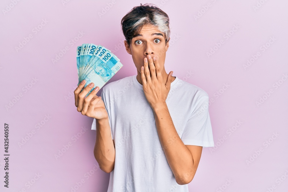 Young hispanic man holding 100 brazilian real banknotes covering mouth with hand, shocked and afraid for mistake. surprised expression Young hispanic man holding 100 brazilian real banknotes covering mouth with hand, shocked and afraid for mistake. surprised expression