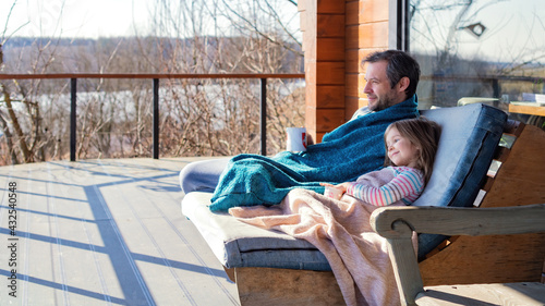 Dad and daughter are sitting together in a chair on the terrace under a woolen blanket. Family vacation out of town concept. Father and daughter are drinking tea, looking at the lake in early spring. #432540548