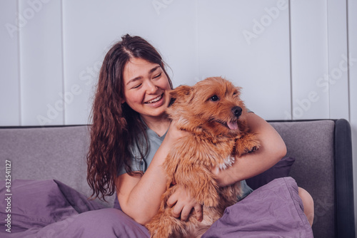 Cheerful smiling woman sitting on the bed hugging her dog Norwich Terrier. Pure friendship concept 