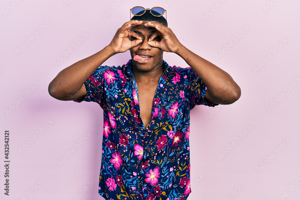 Young black man wearing hawaiian shirt and sunglasses doing ok gesture like binoculars sticking tongue out, eyes looking through fingers. crazy expression.