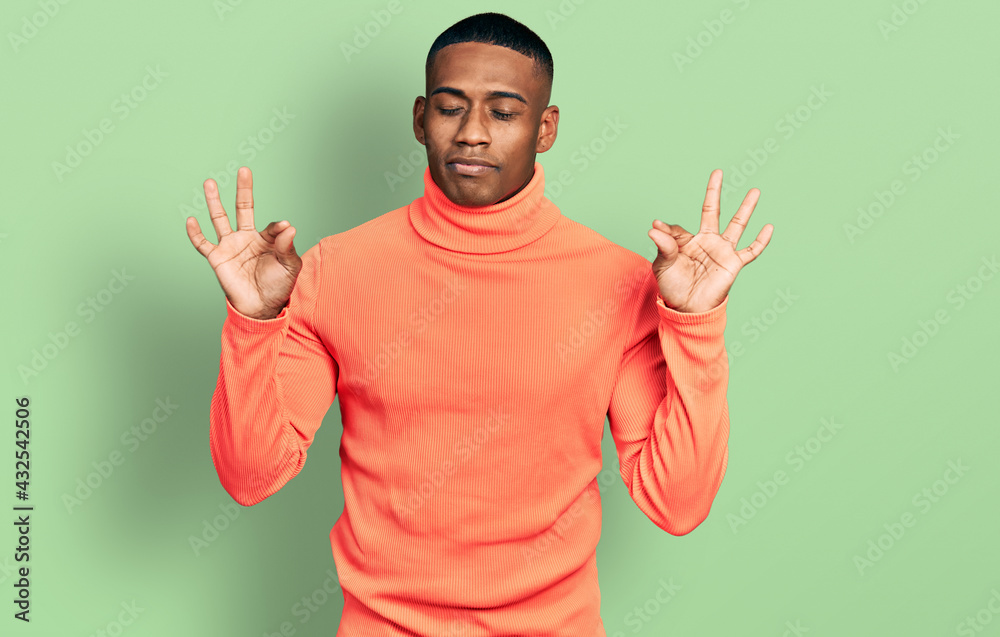 Young black man wearing orange turtleneck sweater relax and smiling with eyes closed doing meditation gesture with fingers. yoga concept. Young black man wearing orange turtleneck sweater relax and smiling with eyes closed doing meditation gesture with fingers. yoga concept.