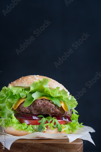Homemade organic hamburger sandwich made of grilled beef ground meat, lettuce, cheese, tomatoes and onion inside sliced bun served on paper on brown wooden table against black background. Vertical