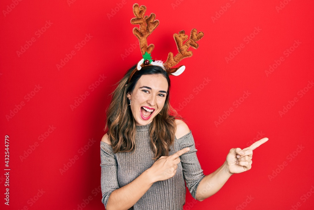 Young hispanic girl wearing deer christmas hat smiling and looking at the camera pointing with two hands and fingers to the side.