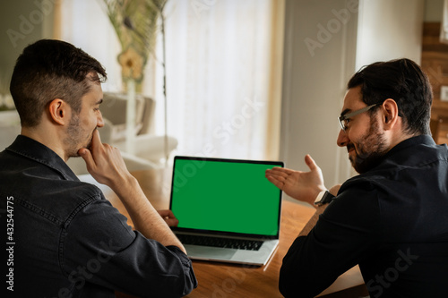 Zwei junge Männer, Anfang 20. sitzen im Wohnzimmer, schauen auf den Display eines Laptops. Einer zeigt mit den Fingern drauf,  der anderer  ist  nachdenklich. Green screen, grüner Bildschirm