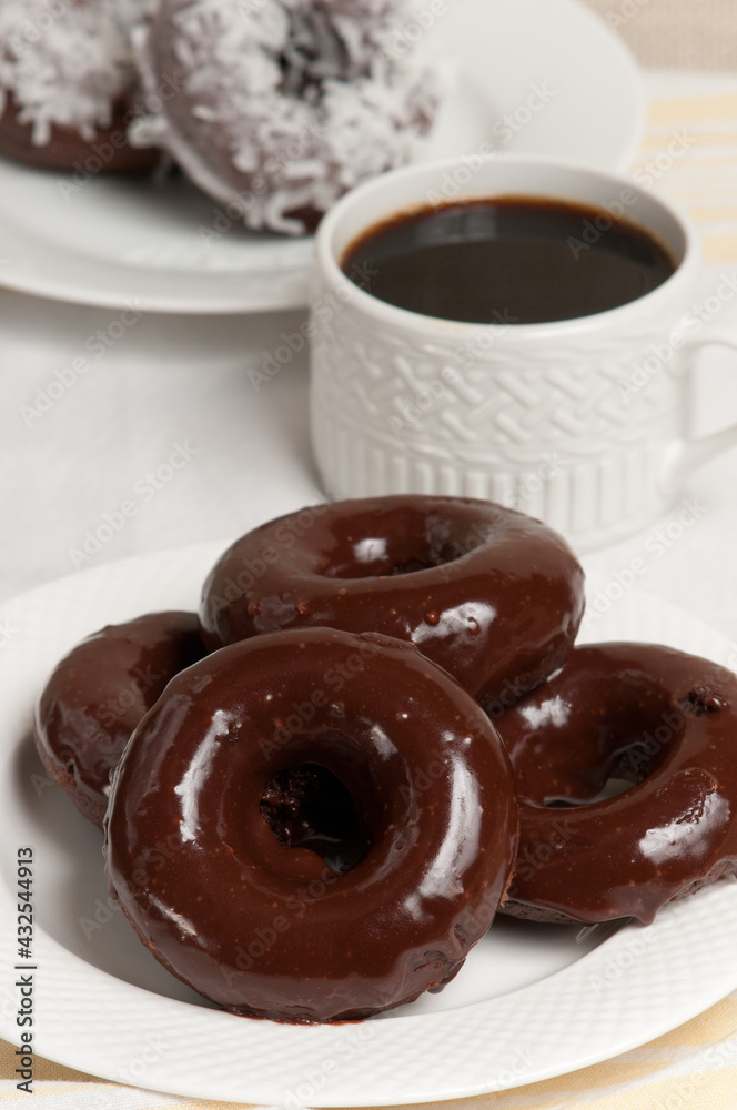 front, top view, close distance, of four freshly backed chocolate donuts covered with dark chocolate frosting on a round, white plate and a cup of black coffee