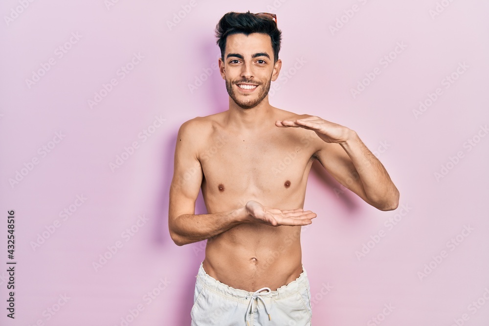 Fototapeta premium Young hispanic man wearing swimwear shirtless gesturing with hands showing big and large size sign, measure symbol. smiling looking at the camera. measuring concept.