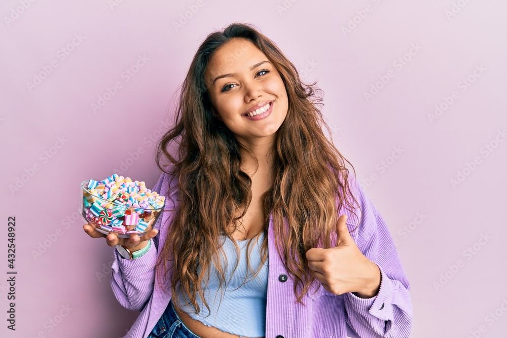 Young hispanic girl holding bowl of sugar candy smiling happy and positive, thumb up doing excellent and approval sign