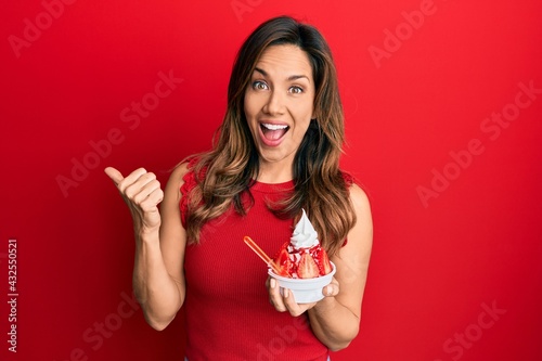 Young latin woman holding ice cream pointing thumb up to the side smiling happy with open mouth