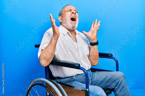 Handsome senior man with beard sitting on wheelchair crazy and mad shouting and yelling with aggressive expression and arms raised. frustration concept. © Krakenimages.com