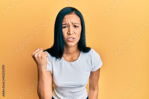 Beautiful hispanic woman wearing casual white tshirt angry and mad raising fist frustrated and furious while shouting with anger. rage and aggressive concept.
