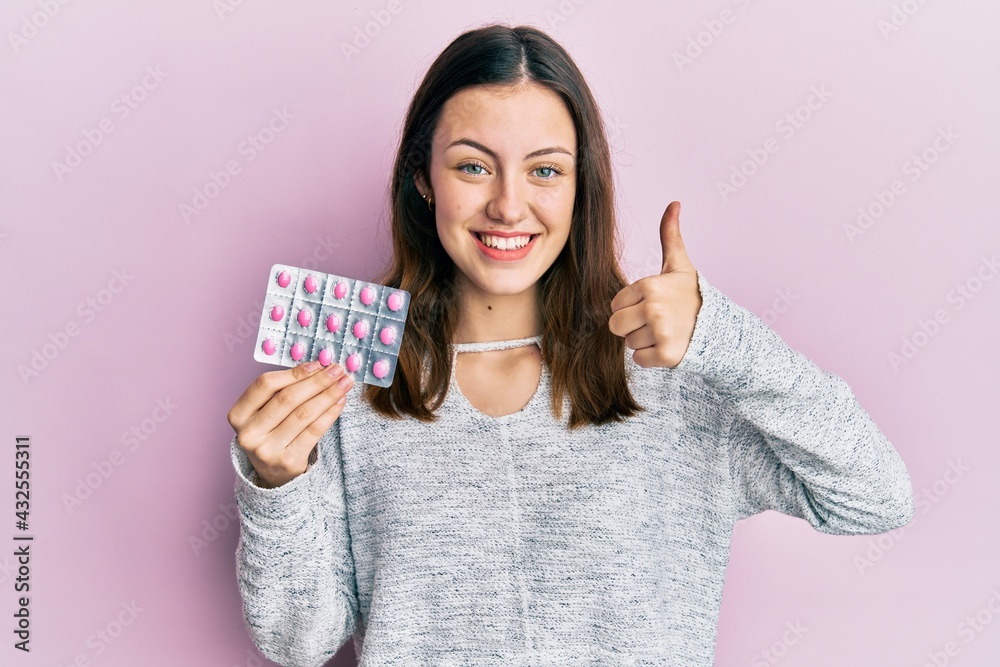 Young brunette woman holding pills smiling happy and positive, thumb up doing excellent and approval sign