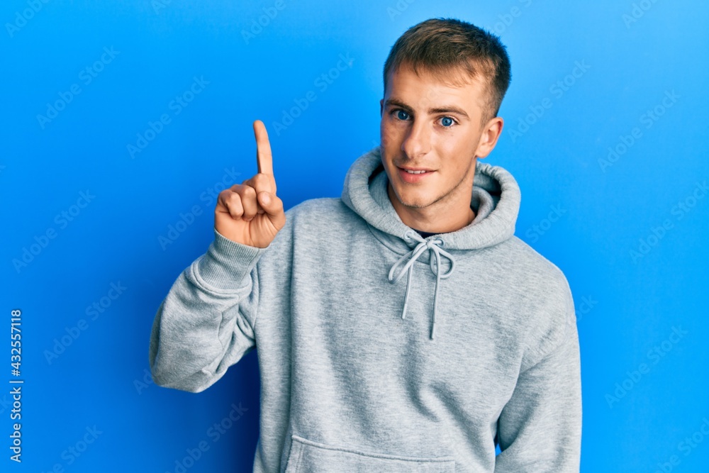 Naklejka premium Young caucasian man wearing casual sweatshirt smiling with an idea or question pointing finger up with happy face, number one
