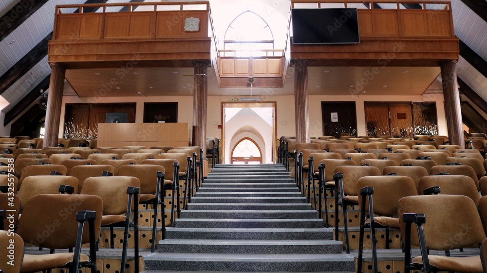 empty hall of a Christian church with a restrained interior in brown tones, rows of soft chairs and a staircase leading up from the bottom with arched windows glowing with daylight