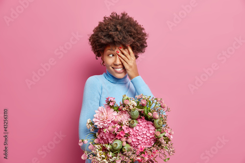 Smiling young African American woman with curly hair covers face and looks away gladfully holds bouquet of flowers received from beloved person enjoys spring time isolated over pink background