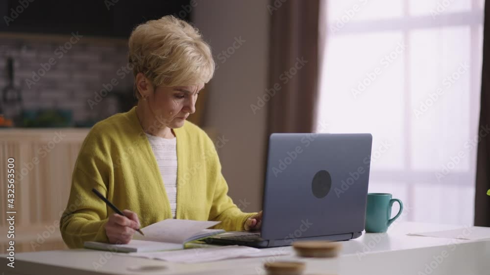 old woman is working remotely at home, using laptop, checking papers and writing in notebook, freelance for accountant and teacher old woman is working remotely at home, using laptop, checking papers and writing in notebook, freelance for accountant and teacher