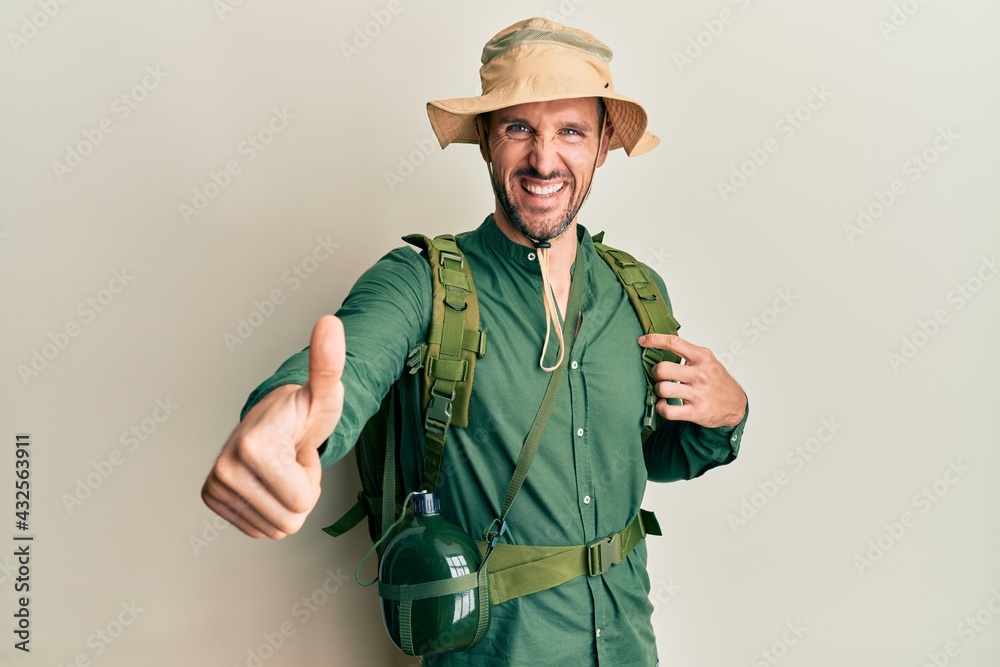 Fototapeta premium Handsome man with beard wearing explorer hat and backpack approving doing positive gesture with hand, thumbs up smiling and happy for success. winner gesture.