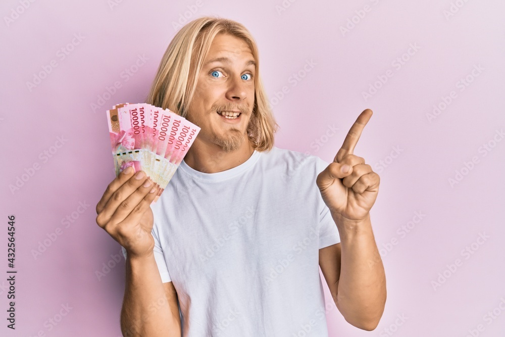 Caucasian young man with long hair holding 100000 indonesian rupiah smiling happy pointing with hand and finger to the side Caucasian young man with long hair holding 100000 indonesian rupiah smiling happy pointing with hand and finger to the side