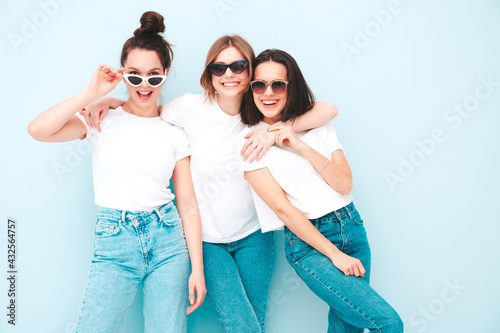 Three young beautiful smiling hipster female in trendy same summer white t-shirt and jeans clothes. Sexy carefree women posing near light blue wall in studio. Cheerful and positive models having fun