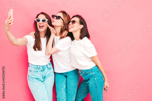 Three young beautiful smiling hipster female in trendy same summer white t-shirt and jeans clothes. Sexy carefree women posing near pink wall in studio. Positive models in sunglasses taking selfie