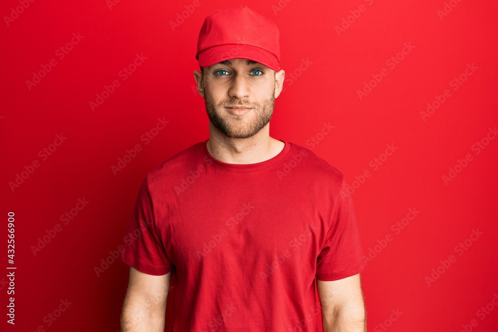 Young caucasian man wearing delivery uniform and cap with serious expression on face. simple and natural looking at the camera.