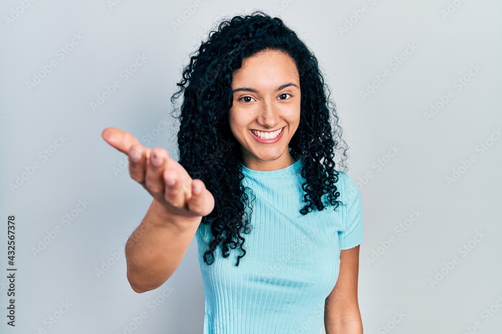Young hispanic woman with curly hair wearing casual blue t shirt smiling friendly offering handshake as greeting and welcoming. successful business. Young hispanic woman with curly hair wearing casual blue t shirt smiling friendly offering handshake as greeting and welcoming. successful business.