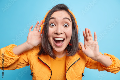 Excited suprised cheerful young Asian woman keeps mouth opened wears headphones on ears listens audio track being shocked and very happy dressed in orange jacket isolated over blue background