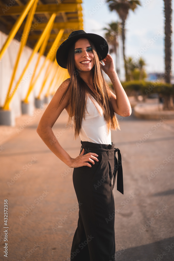 Lifestyle, a young brunette in heels, pants and black hat on a yellow promenade