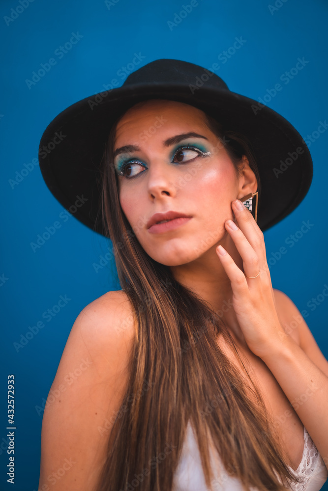 A young brunette in a black hat and white t-shirt looks on a blue background. Blue fashion makeup with sparkles A young brunette in a black hat and white t-shirt looks on a blue background. Blue fashion makeup with sparkles