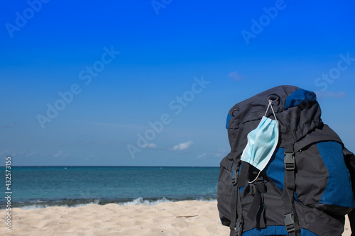A face mask draped over a backpack on a tropical beach signifying the consideration of coronavirus on travel