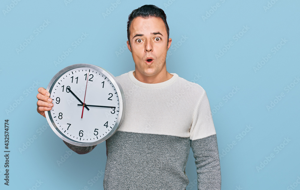 Handsome young man holding big clock scared and amazed with open mouth for surprise, disbelief face Handsome young man holding big clock scared and amazed with open mouth for surprise, disbelief face