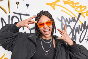 Cheerful cool ethnic woman with dreadlocks makes yo gesture has fun dressed in black jacket and stylish orange sunglasses smiles broadly poses against graffiti background. Youth subculture concept